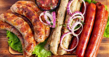 Farklı türde ızgara sosisler ahşap bir tahtada düzenlenir. Salsa, soğan ve marul da dahil. Güneş ışığı yiyeceğin üzerinde parlıyor..