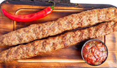 İki uzun kebapçı bir tahta üzerinde bir kase salsa ve kırmızı biberin yanında oturuyor. Sahne yiyeceklerin markette servis edilmeye hazır olduğunu gösteriyor..