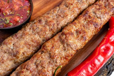 İki kebap, bir kase salsa ve kırmızı biberin yanına ahşap bir tabağa konur. Yemek taze pişmiş ve yenmeye hazır görünüyor..