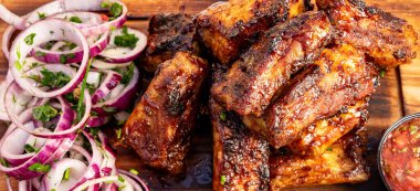 Bir tabak, dilimlenmiş soğan ve taze otların yanında ızgara et parçaları tutar. Yakınlarda bir kase sos var. Mutfak meşgul görünüyor..