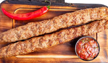 İki uzun kebapçı bir tahta üzerinde bir kase salsa ve kırmızı biberin yanında oturuyor. Sahne yiyeceklerin markette servis edilmeye hazır olduğunu gösteriyor..