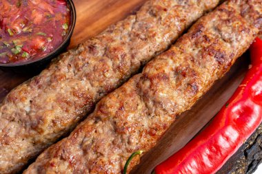 İki kebap, bir kase salsa ve kırmızı biberin yanına ahşap bir tabağa konur. Yemek taze pişmiş ve yenmeye hazır görünüyor..