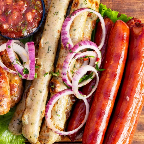 Farklı renkte sosisler tahta bir tahtanın üzerinde. İnce soğan dilimleri üste dizilir. Sosislerin yanında küçük bir kase salsa sosu var..