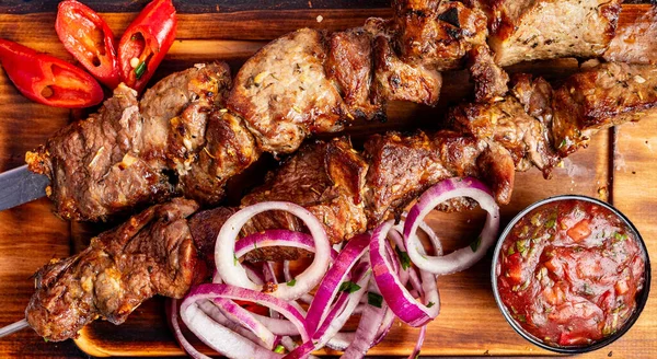 Ahşap tabakta ızgara et şişleri, dilimlenmiş soğanlar, kırmızı biberler ve küçük bir kase sos var..