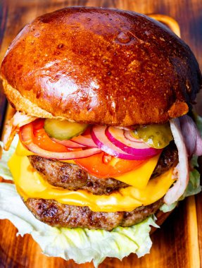 Bir hamburger, iki köfte, çedar peyniri, marul, domates, soğan ve turşu içeren ahşap bir tahtanın üzerinde oturur..
