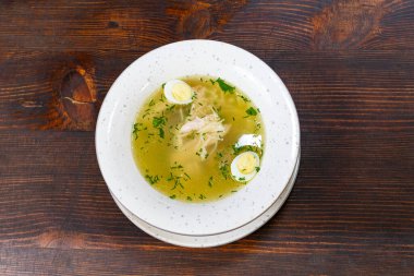Bir kase tavuk çorbası ahşap bir masada oturur. Çorbada tavuk parçaları ve dilimlenmiş yeşil soğan var..