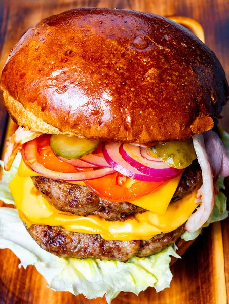 Bir hamburger, iki köfte, çedar peyniri, marul, domates, soğan ve turşu içeren ahşap bir tahtanın üzerinde oturur..