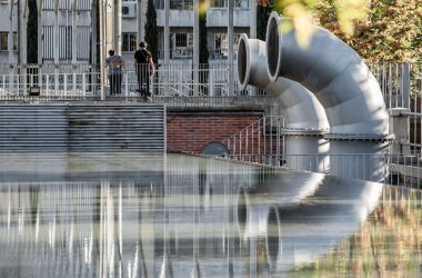 Arka planda iki kadının yürüdüğü iki büyük havalandırma borusunun sudaki yansıması ve endüstriyel bir bina.