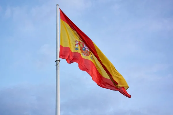 Giant flag of spain hoisted on its mast, on a cloudy day, with blue sky and a lot of wind
