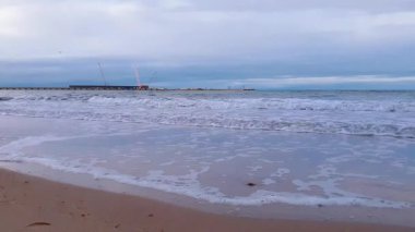 Baltık deniz kıyısındaki ıssız kumsalda kıyı şeridinde fırtına dalgalarının alçak açılı görüntüsü ve bulutlu bir günde bulutlu bir gökyüzünün altında deniz rıhtımı. Hiç kimse sakin deniz manzarası.