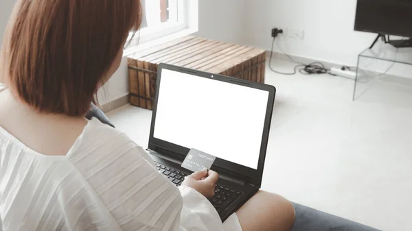 A woman holding a credit card, which is paying online, on a laptop, sitting on the sofa in the living room, at home with clipping path. The concept of online shopping.white screen notebook for mockup