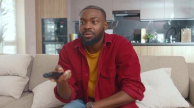 Heyecanlı duygusal Afro-Amerikalı adam televizyonu açıyor. Uzaktan kumandada en sevdiği kanalı seçiyor. Kanepede uzanıyor ve izliyor. Ev eğlencesi kavramı.