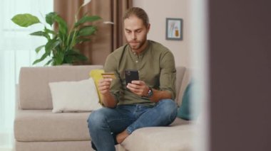 Genç, beyaz bir adam cep telefonu dokunmatik ekranında bankamatik numarasını kullanıyor. Oturma odasında oturmuş online sipariş e-ticaret alışverişi yapıyor..