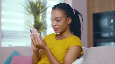 Güzel, çekici afro-amerikan iş kızı cep telefonuyla sosyal medya ağı kuruyor ve kanepede oturuyor. Sıradan insanlar. Ev hayatı. Teknoloji.