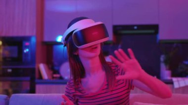 VR gözlüklü mutlu genç kadın evdeki kanepede oturuyor. Neon ışıkta. Hareket eden eller. Teknoloji, oyun yenilikçi eğlence aktivitelerini yenilikçi kılıyor. Yavaş çekim