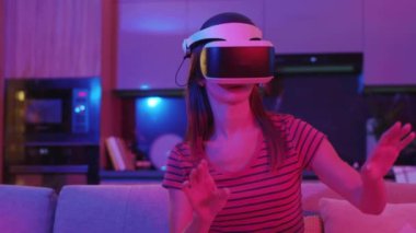 Evde, VR gözlüklü genç bir kadın kanepede oturuyor. Neon ışıkta. Hareket eden eller. Teknoloji, oyun yenilikçi eğlence aktivitelerini yenilikçi kılıyor. Yavaş çekim