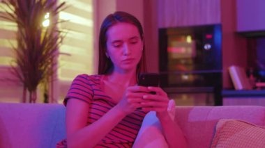 Odaklanmış genç bir kadın, neon ışığın altındaki karanlık odada ciddi bir şekilde telefon dinleyerek sohbet ediyor. Evdeki kanepede dinlenmek. Yavaş çekim