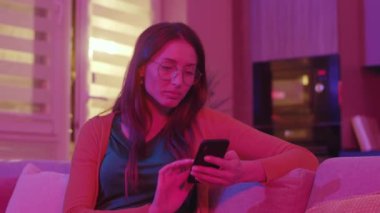 Akıllı telefon ekranına bakan, sosyal medyada sörf yapan, arkadaş paylaşımlarından hoşlanan ciddi sakin genç bir kadın. İşten sonra dinlen. - Eve. Kanepede rahat rahat oturan konsantre bir kız. Neon odası.