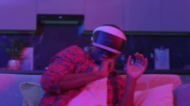 VR kulaklıklı Afro-Amerikalı bir adam sanal bir korku oyunu oynuyor. Evde oturarak el kol hareketi yapan nesneleri aramaktan korkuyor. Neon ışıkları. Oyun eğlencesi. Teknoloji.