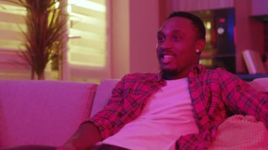 Duygusal Afro-Amerikalı bir adam TV programları izliyor eğlence için kanalları tarıyor. Ev neonu. Aydınlanma. Tasasız yaşam tarzı.