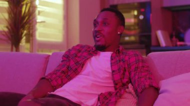 Neon ışığı. Kaygısız, duygusal, Afro-Amerikalı, çizgili tişörtlü, kanepede oturmuş televizyon seyreden adam. Ev eğlencesi ve eğlence konsepti.