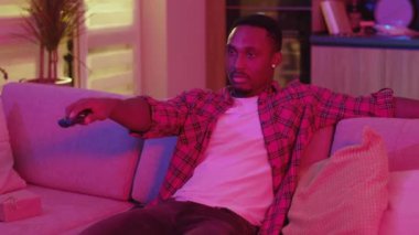 Afro-amerikan çekiciliği olan rahat bir insan kanepe değiştiriyor televizyon seyrediyor uzaktan kumandayla ev eğlencesi neon ışıklarıyla eğleniyor..