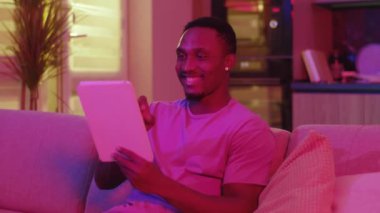 Afro-Amerikalı genç adam tablete göz atıyor internet bağlantısı kuruyor video mesajlarını paylaşıyor evde eğleniyor. Neon aydınlatma. Renkli ev içi.