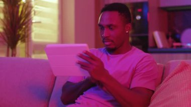 Amerikalı bir adamın dijital tablet bilgisayar teknolojisi aygıtı internet iletişim kullanıcı arayüzüne göz atması ve boş zamanlarını kapalı bir evde geçirmesi. Neon konsepti.
