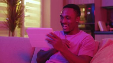 Duygusal neşe saçan Afrikalı adam tableti karıştırıyor uzaktan haberleşme video kaseti yayınlamak için uzaktaki arkadaşını arıyor oturma odasının neon ışıklarında kalıyor..