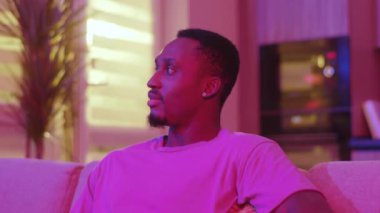 Yakışıklı, çekici, sıradan, modern Afro-Amerikan bir adamın portresi. Kameraya gülümsüyor. Renkli neon renkli oturma odasında dinleniyor..