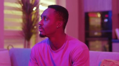 Çekici modern samimi Afro-Amerikan yakışıklı genç adam oturma odasında dinlenirken gülümsüyor şık neon ışıklar renkli ev ışıkları.