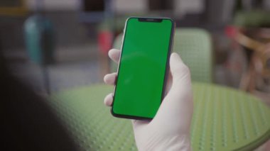 Genç adam boş restoranda oturuyor, cep telefonu kullanıyor. Arka planda bulanık yeşil masa. Yüzü olmadan. Beyaz tıbbi eldivenli ellerin yakın çekimi. Yemek molası, karantina zamanı. Kapalı.
