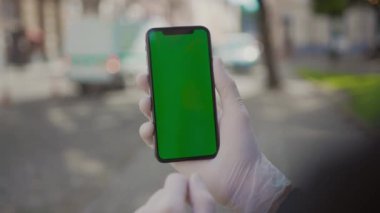 Akıllı telefonu elinde tutan genç adam bulanık sokak arka planındaki ekranı işaret ediyor. İnternetten alışveriş, internet pazarlaması. Eldivenli adam dışarıda cep telefonu kullanıyor. İzole edilmiş, yakın plan.