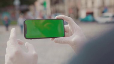 Tıbbi koruyucu eldivenli adamın yakın görüntüsü. Genç Avrupalı adam akıllı telefonu elinde tutuyor, sosyal ağlarda sörf yapıyor, sohbet ediyor. Yüzü olmadan. Karantina zamanı, covid-19 konsepti, fikir. Şehir