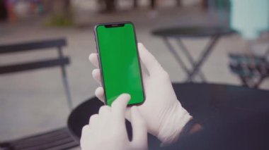 Beyaz koruyucu eldivenlerle siyah masa ve kafein sandalyelerinin bulanık arka planında akıllı telefon kullanan erkek eller. Terasta oturan genç iş adamı yeşil ekran cep telefonuna dokunuyor..