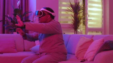 Komik gülümseyen melez genç adam evdeki koltukta oturan sanal gerçeklik kulaklığını çıkarıyor. Mutlu neon ışığı hisset. Eğlenceli oyundu. VR gözlükleri. Yavaş çekim