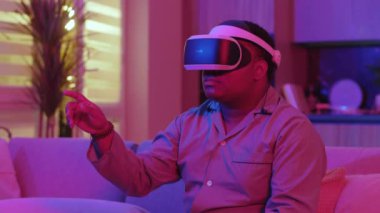 Kanepede oturmuş sanal gerçeklik kulaklığı takan ciddi melez genç adam evde neon ışığı altında parmağını oynatmayı seçiyor. Eğlenceli oyundu. VR gözlükleri sosyal ağ robotu. Yavaş çekim