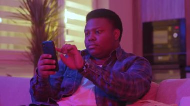 Ciddi bir Afrikalı genç Amerikalı mesaj yazıyor ve akıllı telefon ekranına bakıyor. Kanepede oturup evdeki sosyal ağlarda sohbet etmek. Yavaş çekim