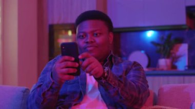Gülümseyen genç Afrikalı Amerikalı adam mesaj yazıyor ve akıllı telefon ekranına bakıyor. Kanepede oturup evdeki sosyal ağlarda sohbet etmek. Yavaş çekim
