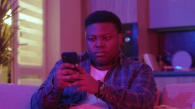 Portre, mesaj yazan ve akıllı telefon ekranına bakan Afro-Amerikan genç bir adama odaklandı. Kanepede oturup evdeki sosyal ağlarda sohbet etmek. Yavaş çekim