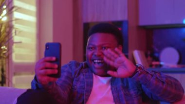 Neşeli, Afro-Amerikalı genç bir adam akıllı telefon ekranına bakarak görüntülü konuşma yapıyor. Kanepede oturup evdeki sosyal ağlarda sohbet etmek. Yavaş çekim