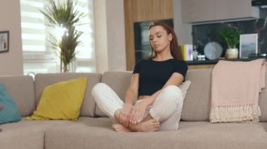 Genç ve güzel bir kadın kanepede oturuyor ve evde gözleri kapalı meditasyon yapıyor. Gülümseyerek mutlu ol. İyi bir denge dengesi. Yavaş çekim