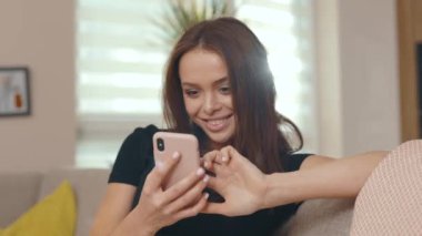 Kanepede oturan ve modern odada akıllı telefon kullanan mutlu genç bir kadın. Güneşli odada gülümsüyor. İnternet sosyal medya bağlantısı. Yavaş çekim