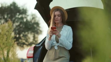 Güzel görünümlü, şapkalı, arabanın yanında duran ve akıllı telefon kullanan beyaz bir kadın. Sosyal medyada izole edilmiş gülümseyen kız mesajları. Arka planda bulanık doğa. Hafta sonu açık hava
