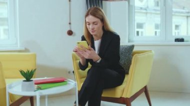 Sosyal ağları kontrol eden başarılı bir halkla ilişkiler müdürü. Sarı koltukta oturan ve ofisinde akıllı telefon kullanan siyah takım elbiseli, sarışın, beyaz bir kadın. İçeride. Dijital