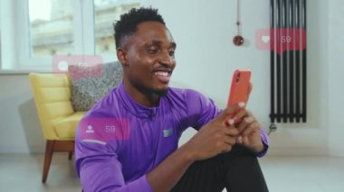 Mor spor ceketli mutlu bir Afrikalı dairede akıllı telefondan internete bakıyor. Gülümseyen spor koçu sağlıklı yaşam tarzı hakkında blog yazıyor, abonelerle oturma odasında iletişim kuruyor...