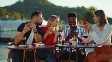 Birbirine aşık iki çift kafeteryada oturmuş akıllı telefonlara bakıyor. Afrikalı adam sosyal ağlardan arkadaşlarına resimler gösteriyor. Gençler konuşuyor, gülüyor. Açık havada. Çok kültürlü insanlar.