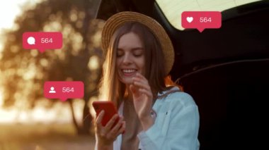 Şapkalı, arabanın bagajında oturan ve akıllı telefon kullanan genç, pozitif bir kadın. Gülümseyen muhteşem kızın beyaz gömlek giydiği görüntüler. Doğal güzellik. Sonbahar geldi. Arka plan bulanık. Açık havada. Sohbet