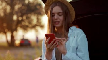 Beyaz tenli güzel bir öğrencinin dışarıda dinlenirken çekilmiş bir portre görüntüsü. Akıllı telefon kullanıyor, yeni fotoğraflar yüklüyor. Şık giyinmiş genç bir kadın mesaj atıyor, gülüyor, başka tarafa bakıyor. Hareket. Bulanık arkaplan