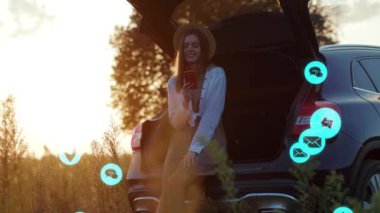 Şapkalı genç ve güzel bir kadın araba bagajında otururken akıllı telefon kullanıyor. Vlog tutucu sosyal medya, uygulama simgeleri mavi arayüzlü yüksek sesle canlandırma. Yavaş çekim